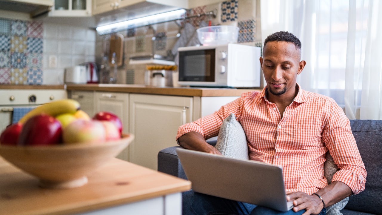 Man sitting on couch with laptop at home smiling, kitchen in background