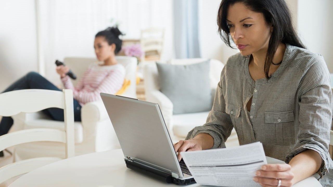 A woman looks over her finances on a laptop and on paper while a teenage girl sits in the background.