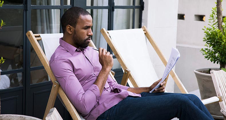 Man in purple shirt reading paperwork in patio chair | PhotoAlto/Sigrid Olsson/Getty Images