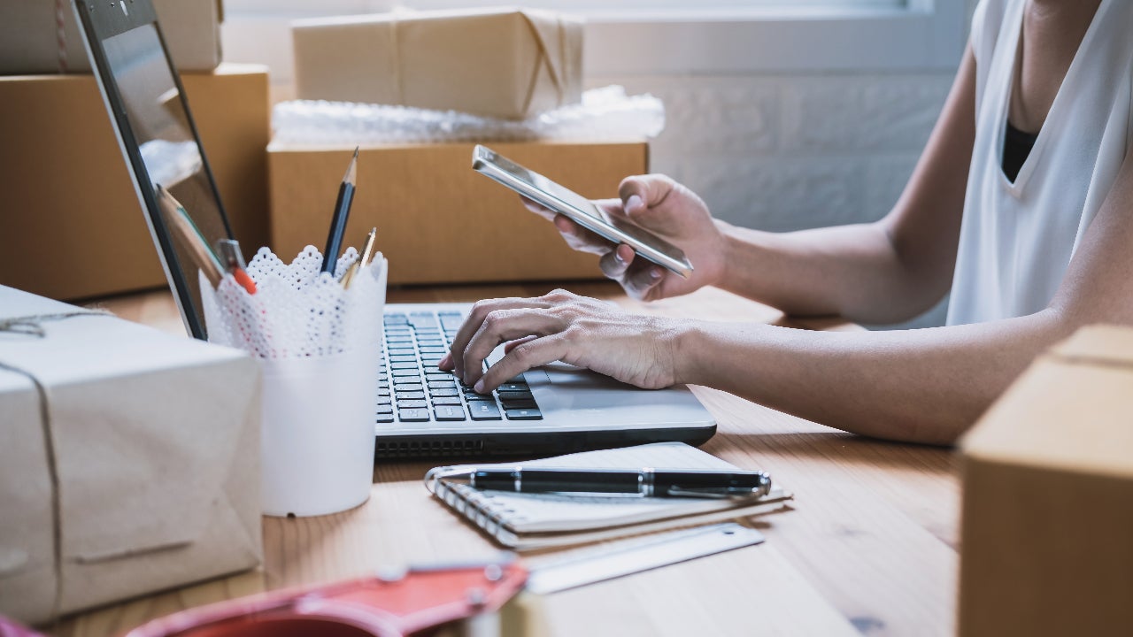 person with laptop looking at cell phone, surrounded by boxes