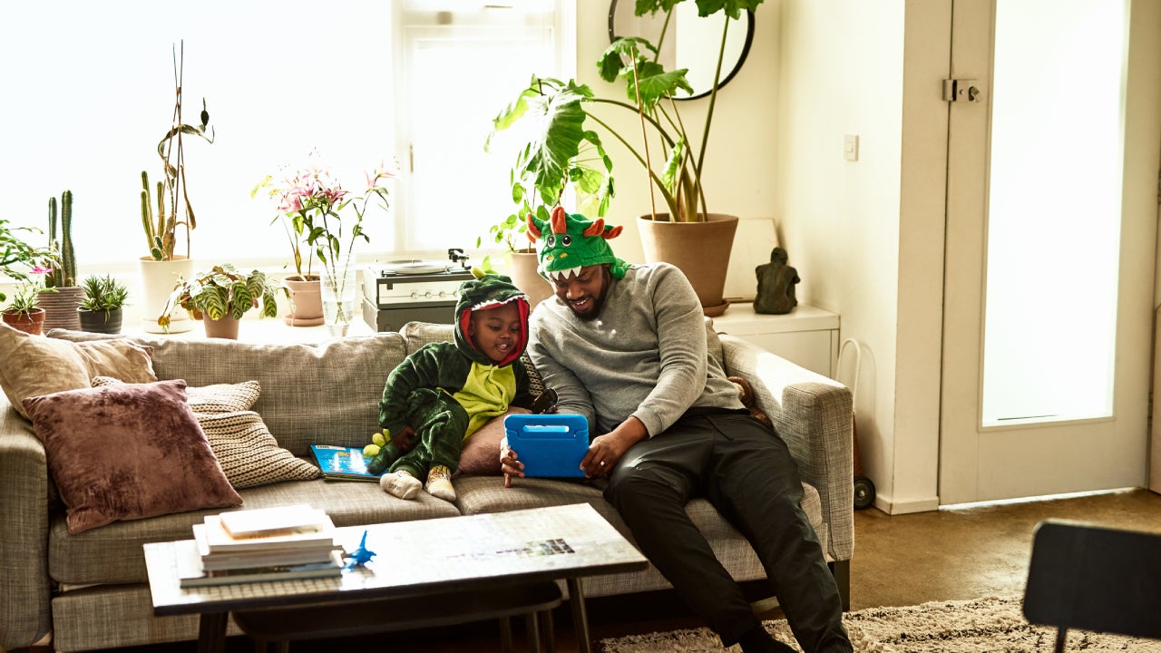 A father and his son, dressed up like dragons, do a learning exercise in the living room.