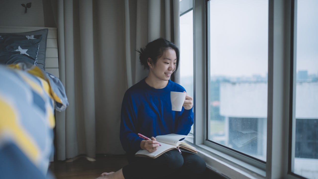 Uma estudante asiática sentada junto à janela do seu apartamento olhando para a cidade com uma caneta e um bloco de notas numa mão e uma chávena de café na outra.