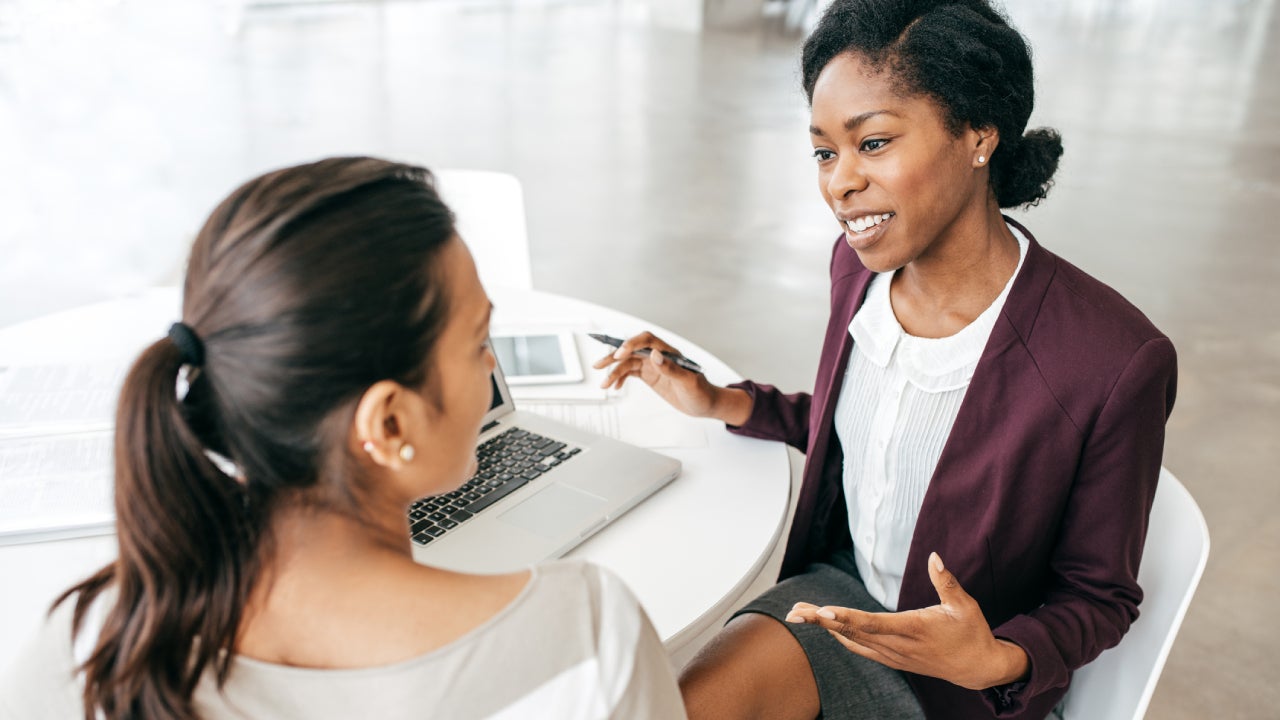 A black lady who is a financial advisor is sitting down on a white table across from a Caucasian woman client. They are talking about something and a laptop is open in the table.