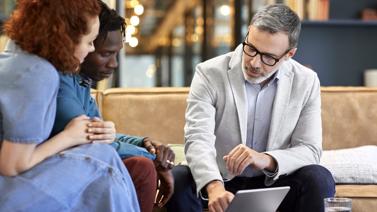 Interracial couple sits on a couch across from a financial expert to review a policy.