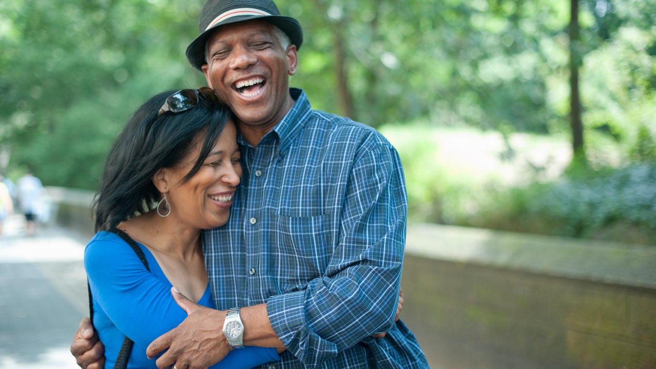 A middle-aged Black couple holds one another laughing in front of a park