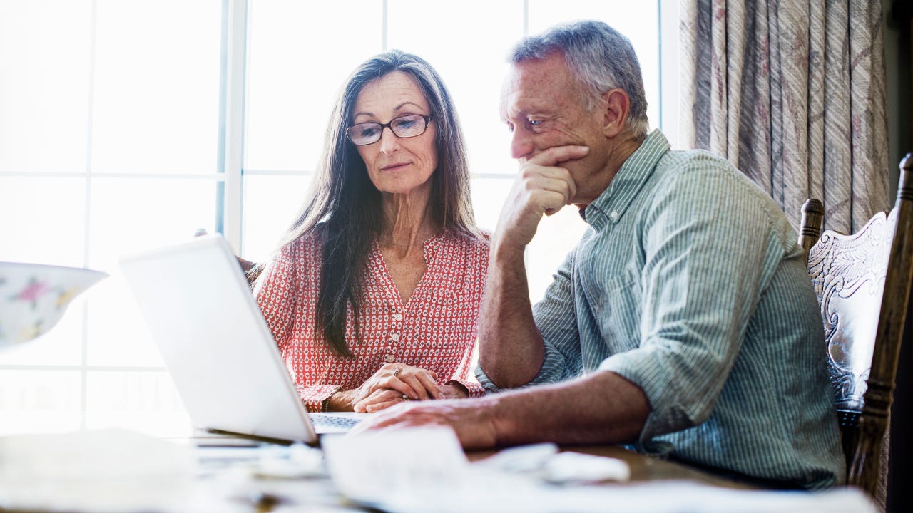 Senior couple sitting at a dining table, using a laptop computer, paperwork and bills on the table