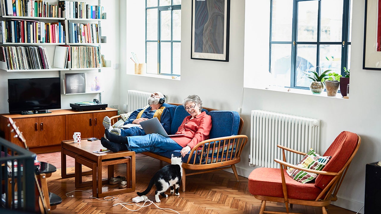 Retired couple sitting on sofa at home, relaxing, woman with laptop smiling, man with head back wearing headphones, lost in music, sitting side by side on blue sofa in retro style living room