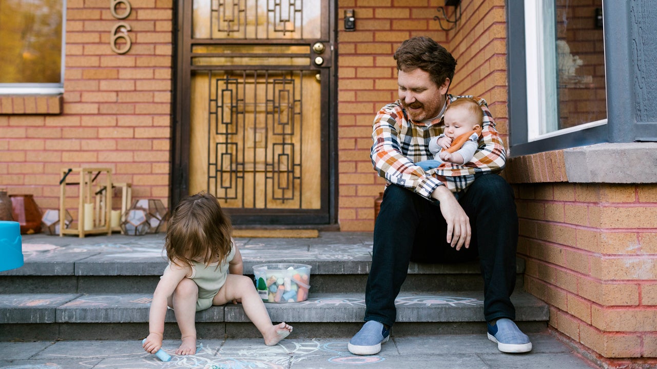 A parent sits on the stoop in front of their home with one baby child in their arms and another, toddler, drawing with chalk on the walkway.