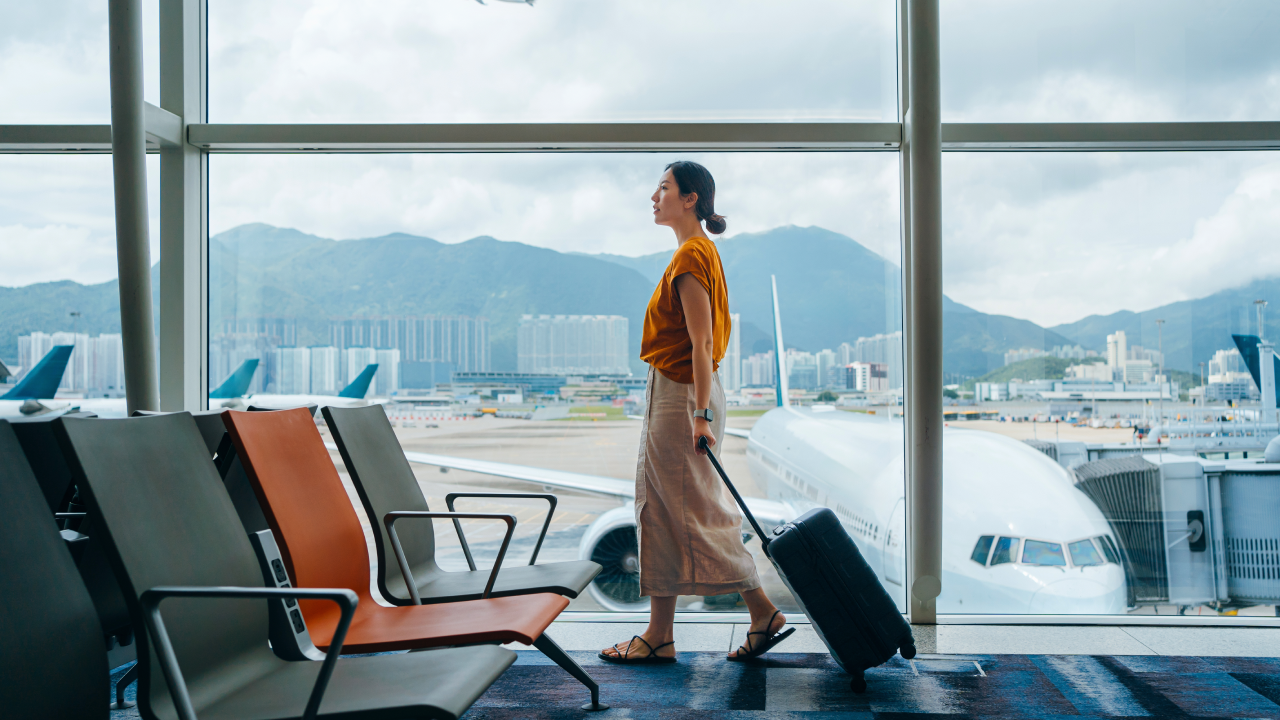 Young Asian woman carrying suitcase, walking by the window at airport terminal. Young Asian female traveller waiting for boarding at airport. Business travel. Travel and vacation concept