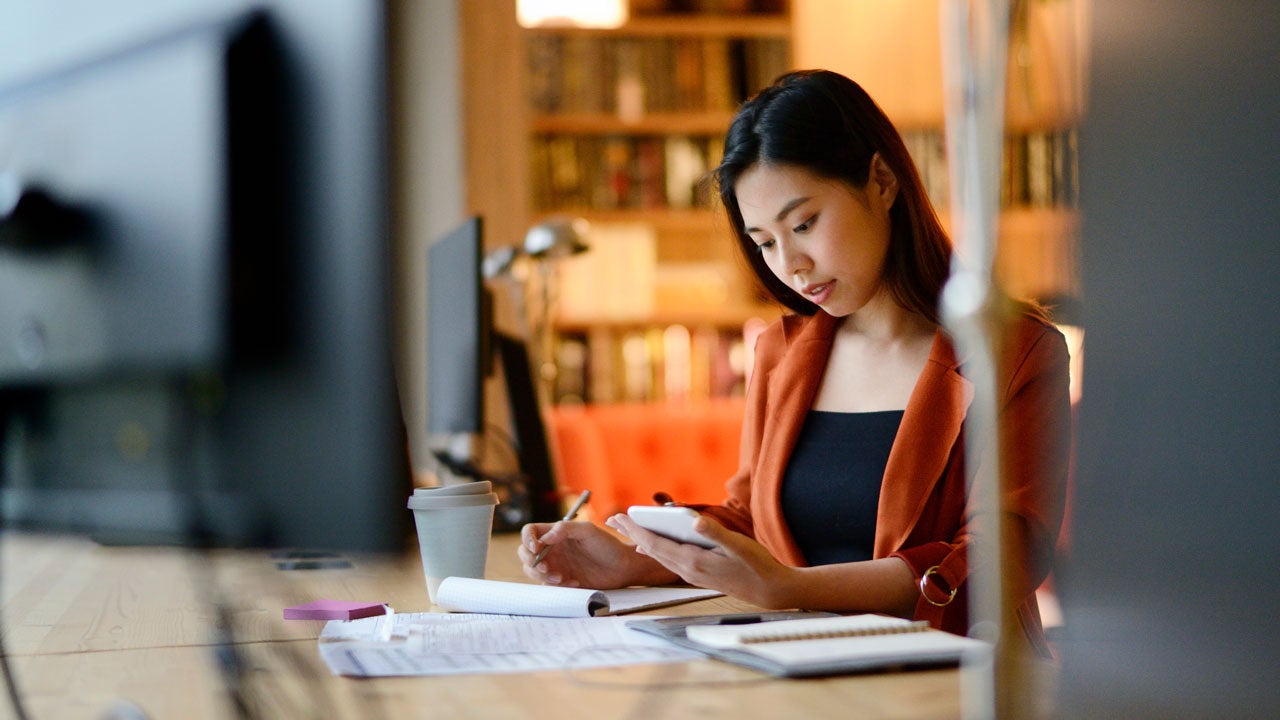 woman writing in notebook and looking at her phone