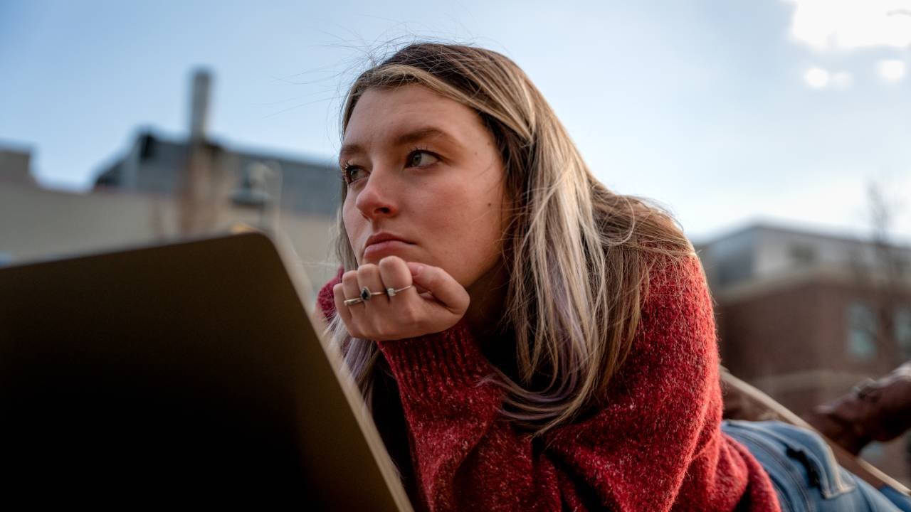 A Young University Student Laying On The Lawn Looking At Her Computer, Deep In Thought