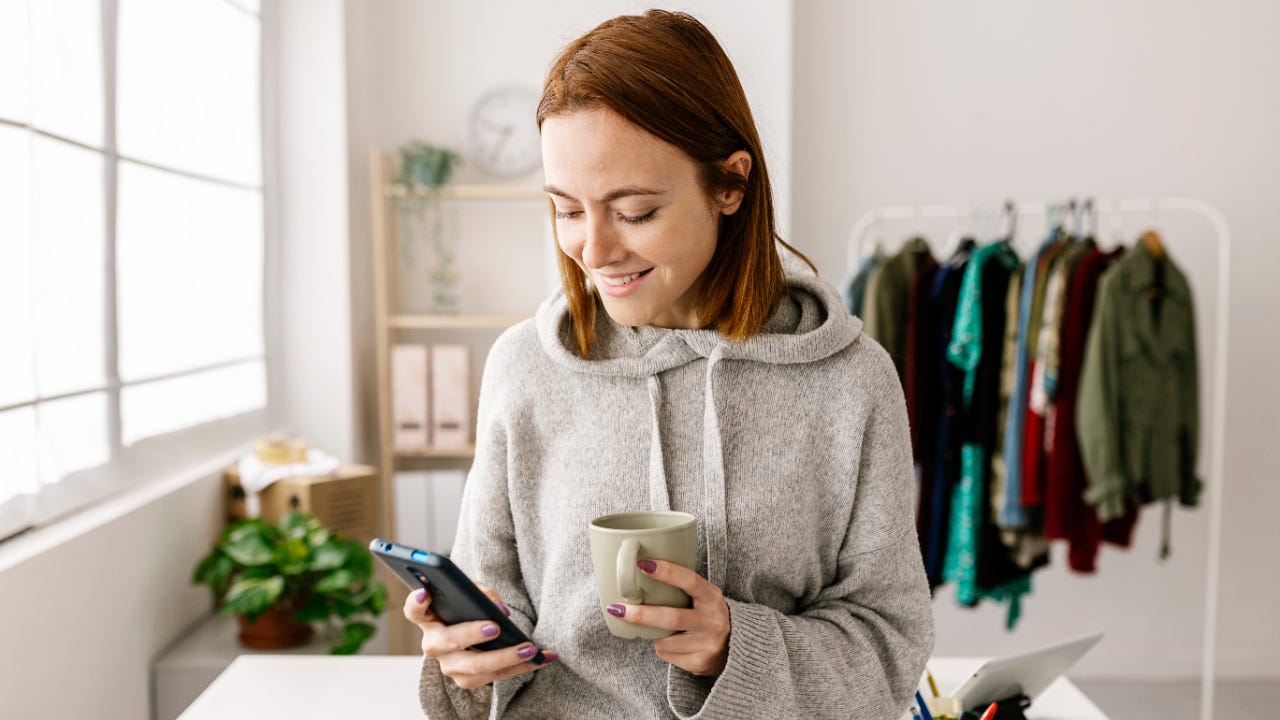 Woman holding coffee in front of desk and clothes stand, checking her phone and smiling