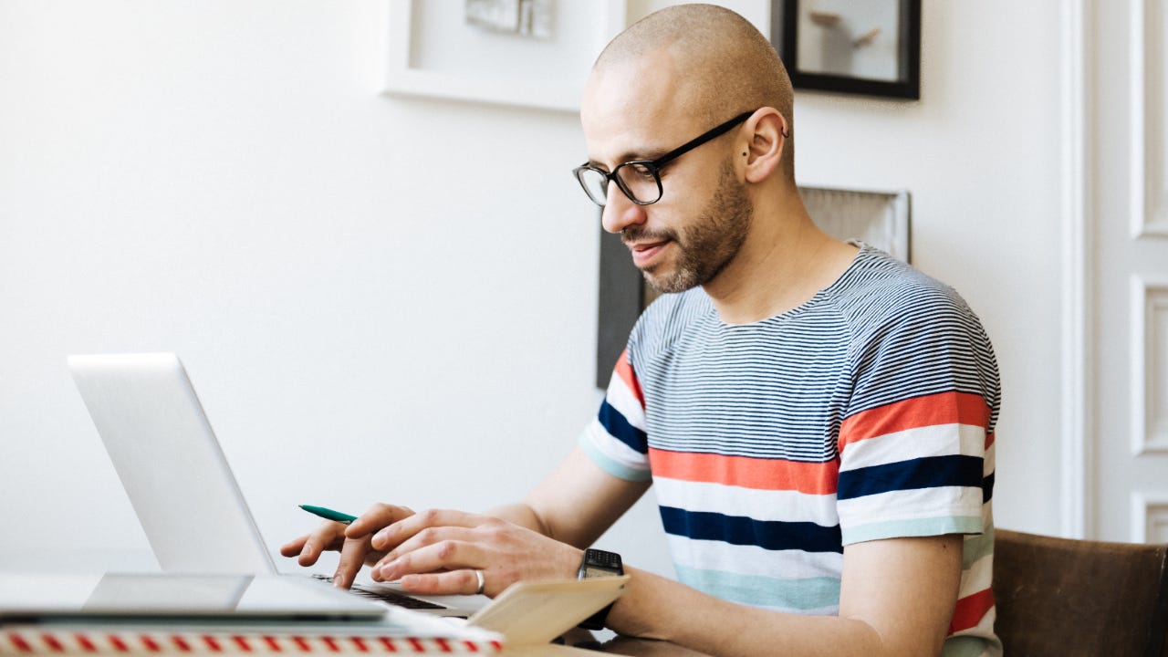 Man in a striped shirt smiling and typing on laptop at home table