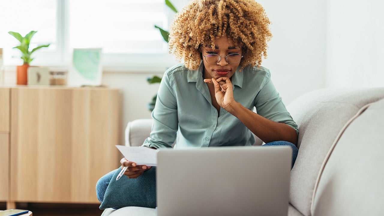 Serious woman is sitting on a sofa and looking at a laptop while holding a paper in her hand. She is wearing glasses. She might be working from home or studying, or paying a utility bill. She is wearing a green shirt and jeans.