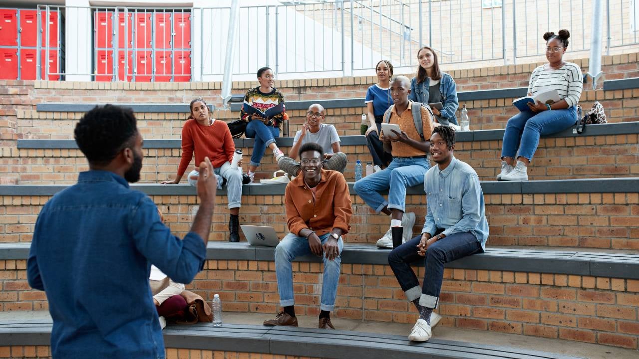 Young male professor teaching multi-ethnic students sitting on amphitheater steps at university campus