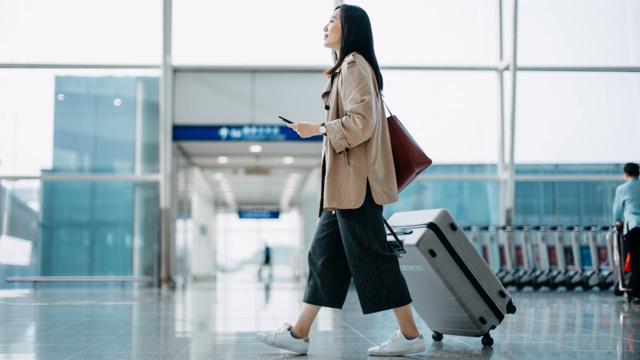 Young woman carrying suitcase and holding smartphone in hand, walking in airport terminal.