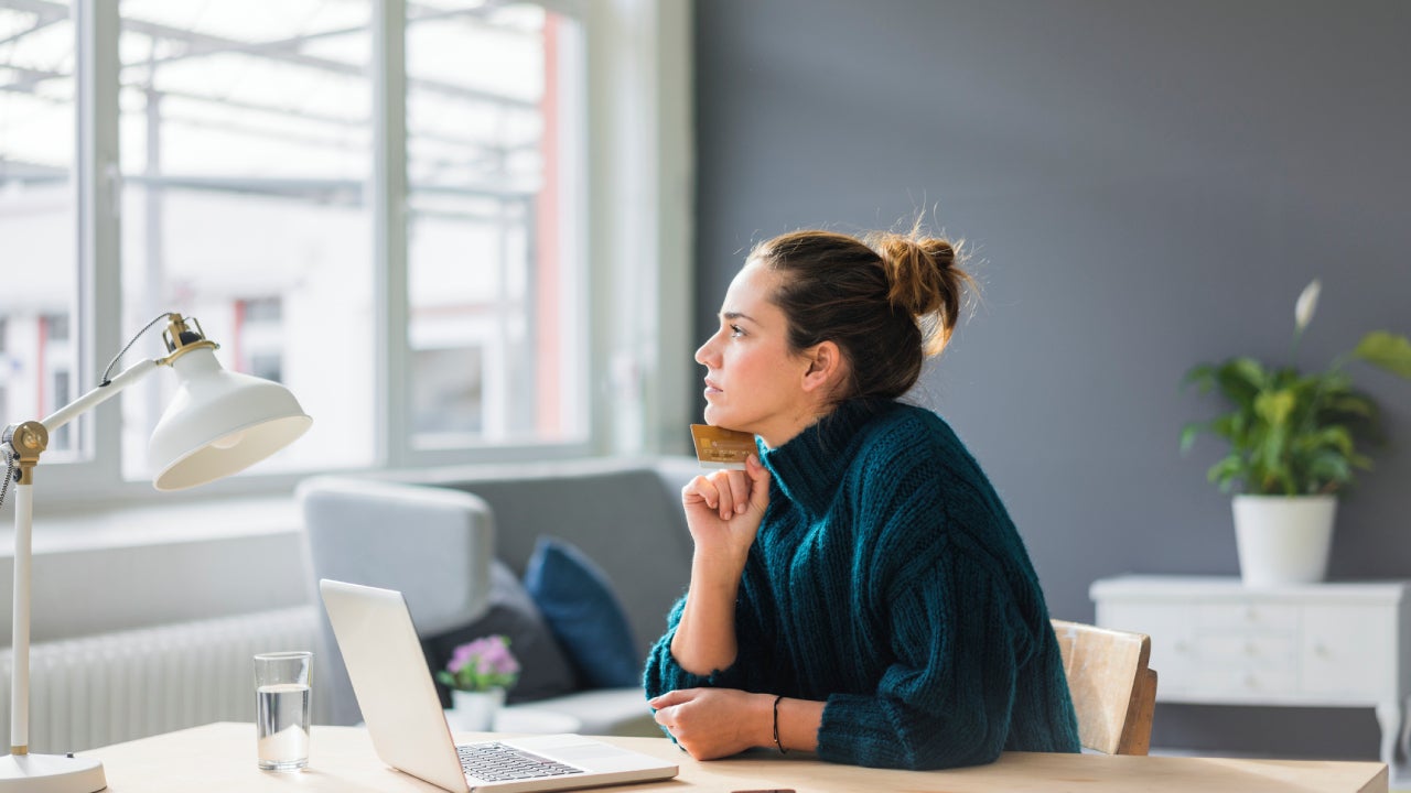 Profile of pensive woman with laptop and credit card sitting at desk at home looking out of window