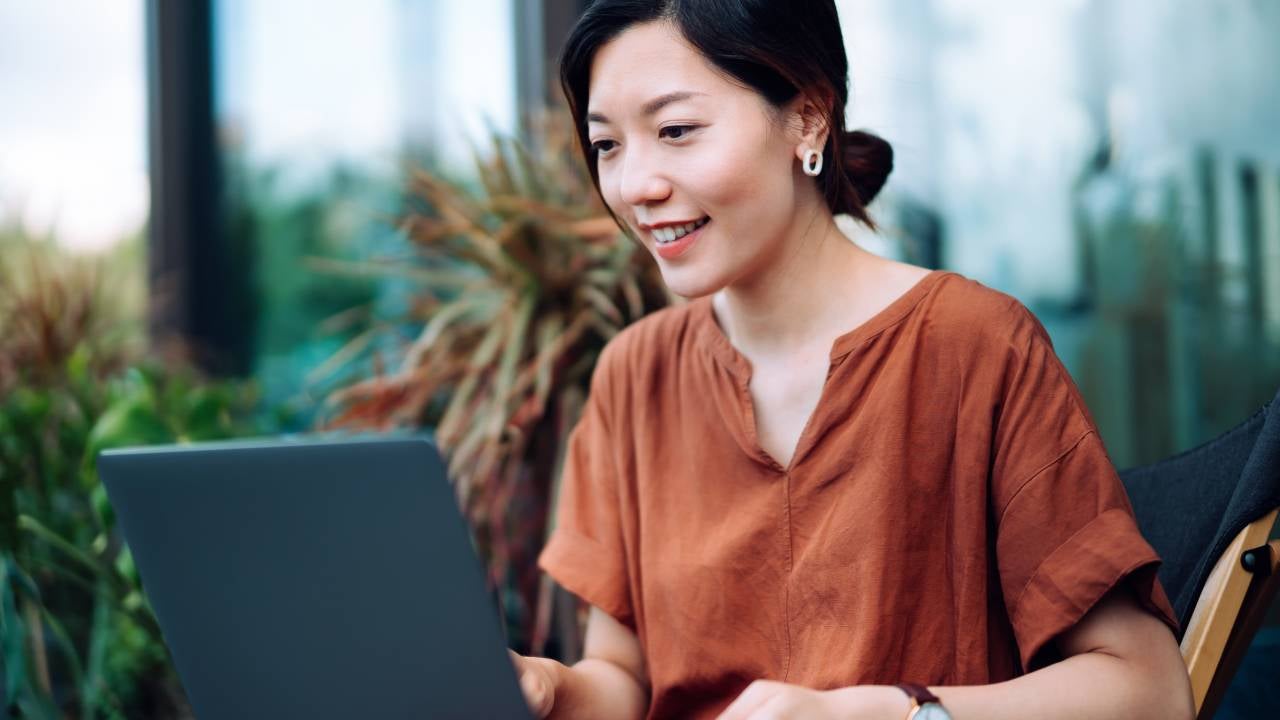 Beautiful young Asian woman sitting on deck chair in the balcony, managing banking and finance with online banking on laptop.