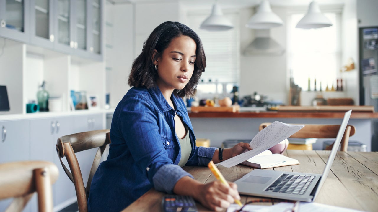 Shot of a young woman using a laptop and going through paperwork