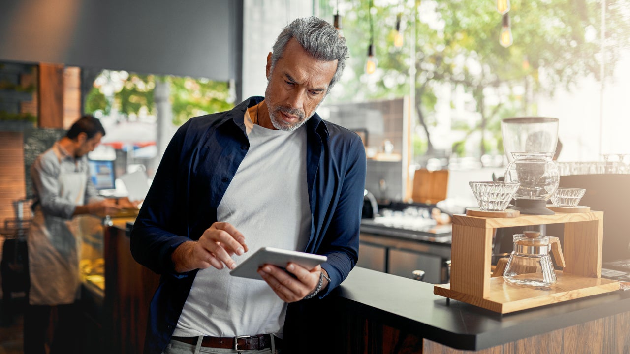 A coffee shop owner leans on the counter, looking at his laptop while a barista works in the background.