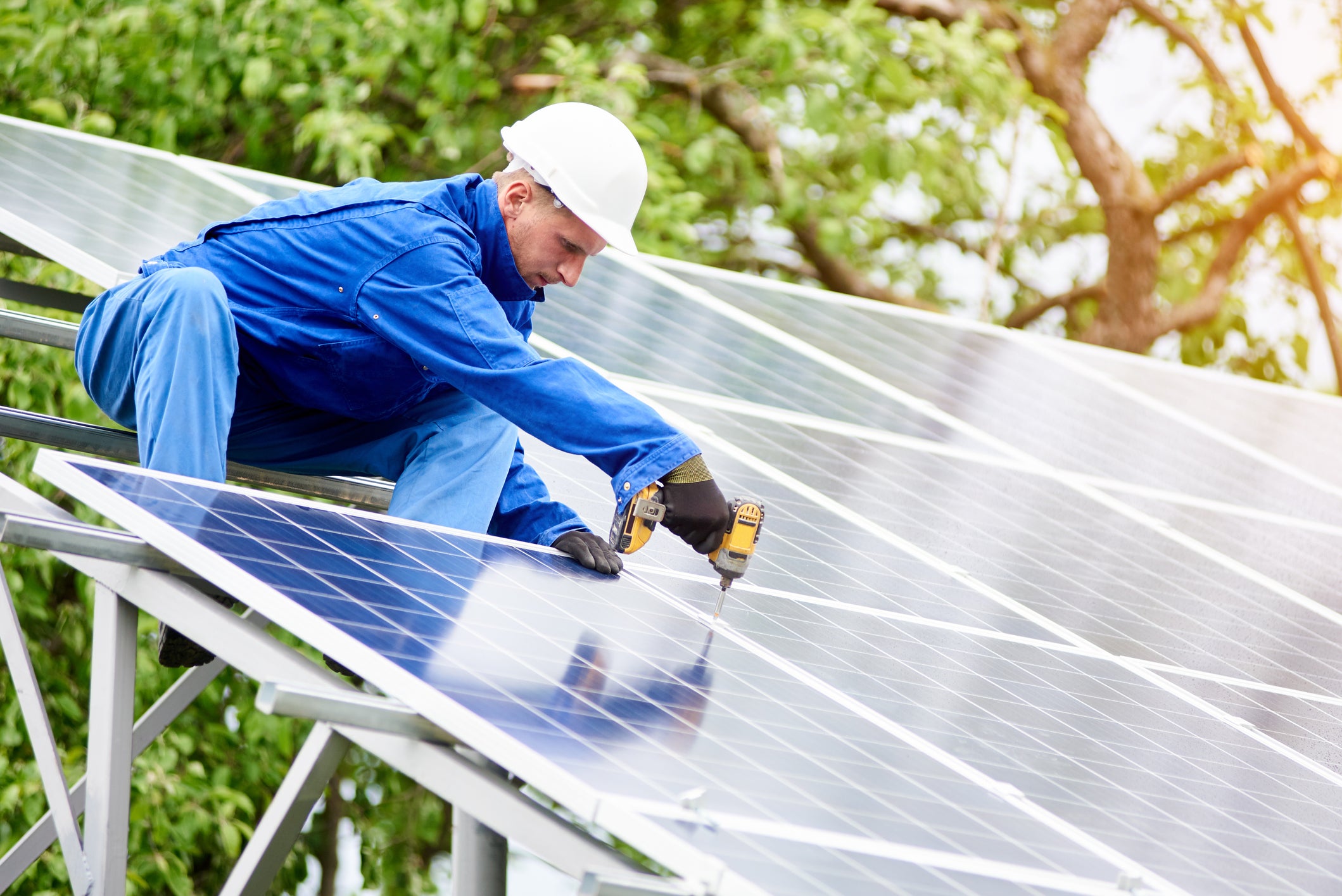 Young construction worker connects photo voltaic panel to solar system using screwdriver on bright sunny day.