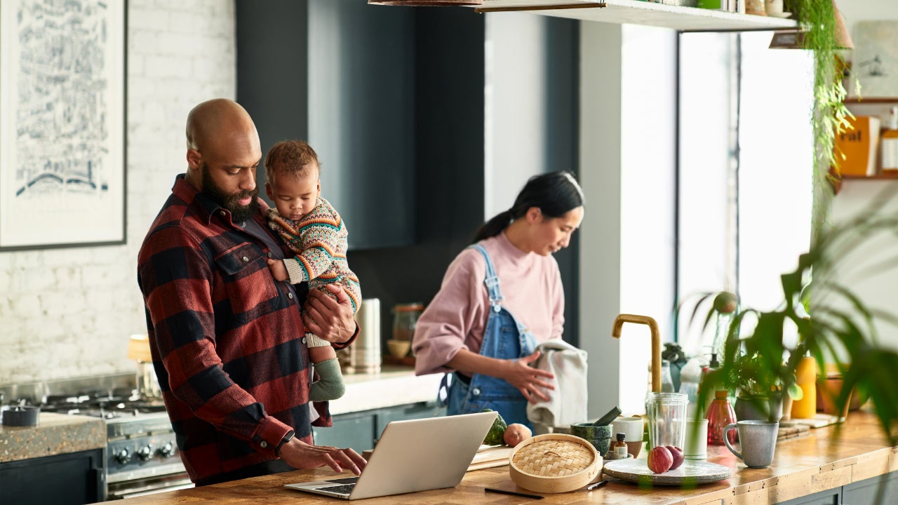 A mother prepares a meal in the kitchen as father holds baby boy and uses a laptop.