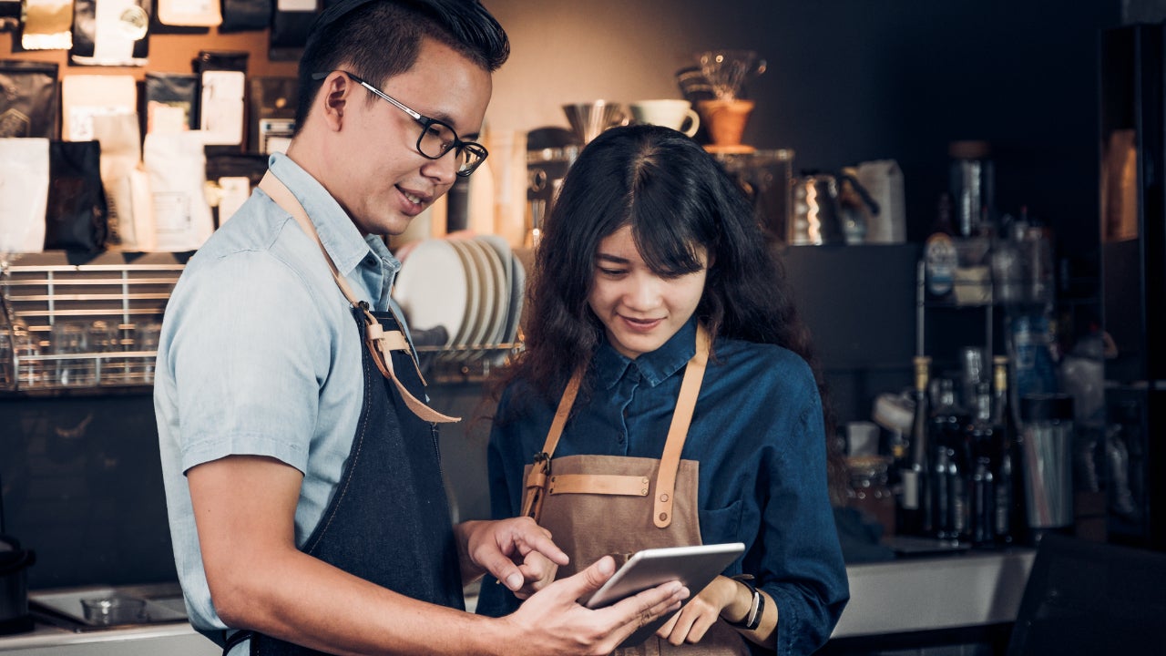 Two coffeeshop owners in aprons stand and look at a tablet in their kitchen.