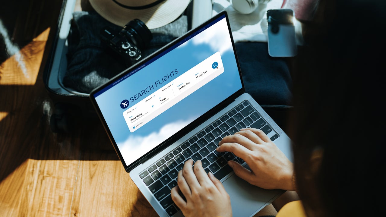 Young Asian woman shopping online for flight ticket on airline website with laptop at home, sitting next to suitcase with sun hat, camera, clothings, headphones, smartphone and passport. Getting ready for vacation. Travel and vacation concept