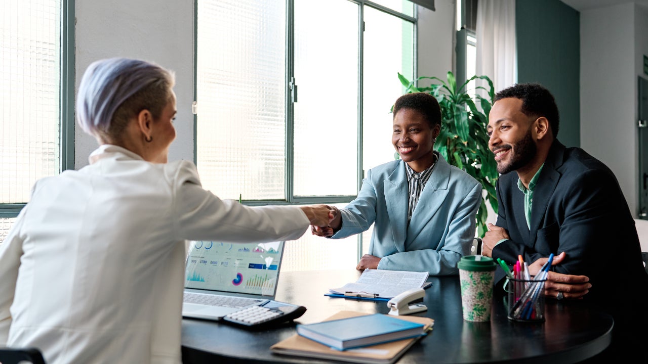 A lender shakes the hand of a smiling business owner who sits next to her partner in the lender’s office.