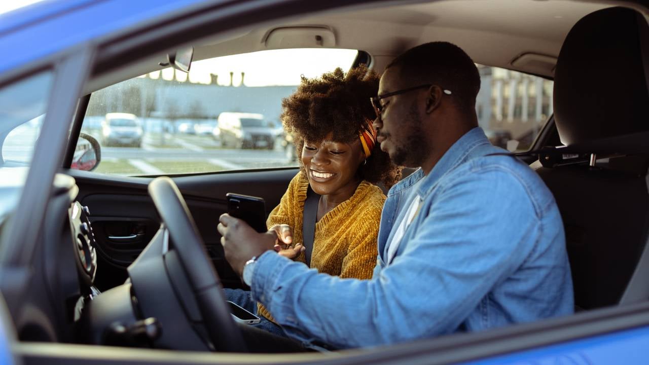 Cheerful Black man and woman using smartphone in a parked car.