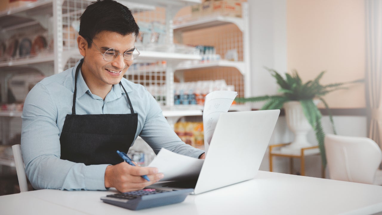 Male business owner in glasses using a laptop while reviewing documents.