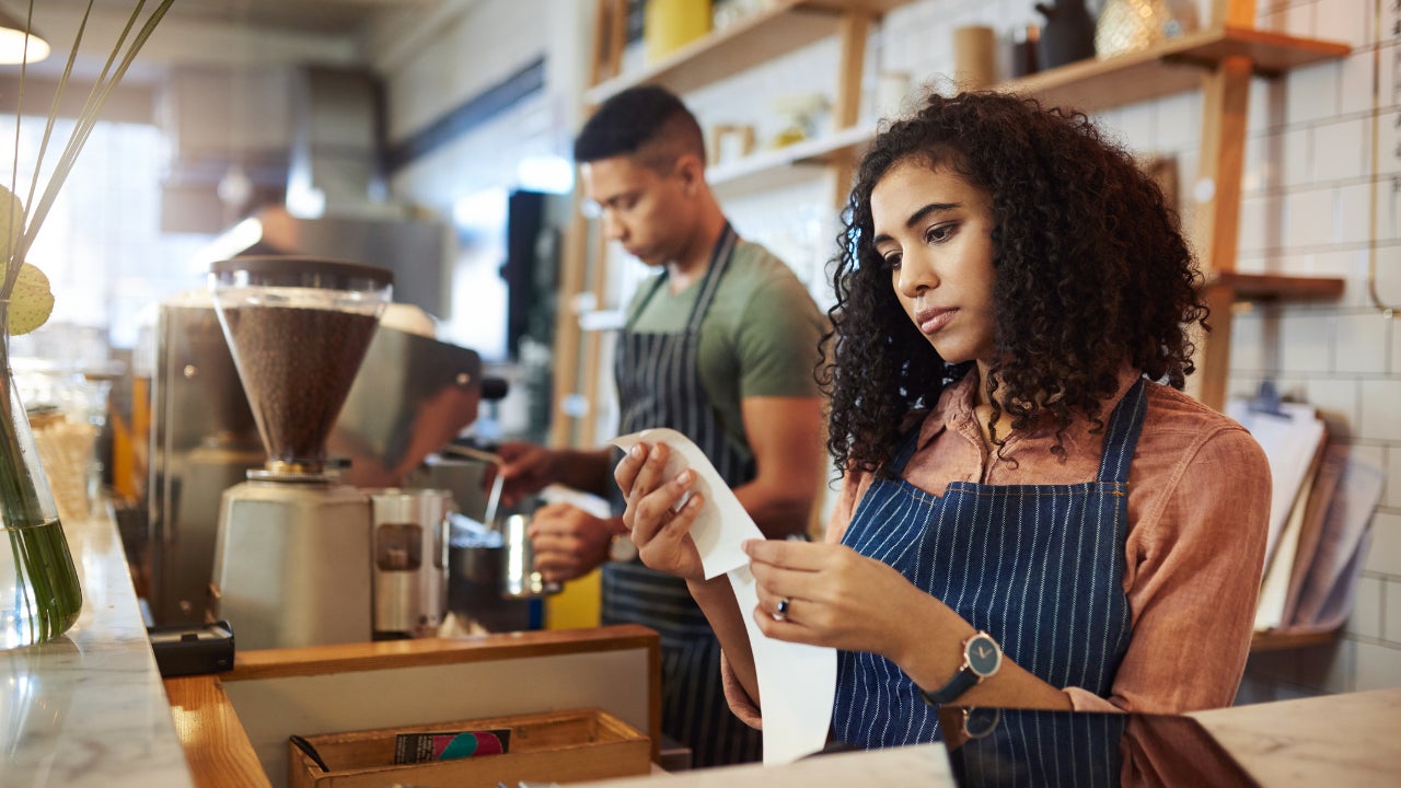 A young business owner stands behind the counter of her coffee shop, looking discouragingly at the day’s receipts.