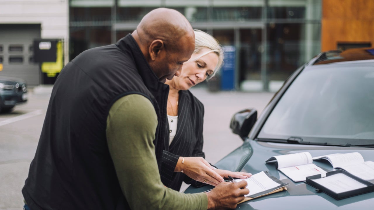 Saleswoman assisting male customer while doing paperwork standing near car