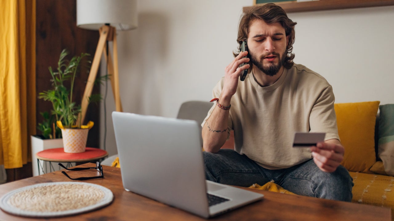 Man is sitting on a sofa in the living room, holding a credit card in his hands and making a phone call