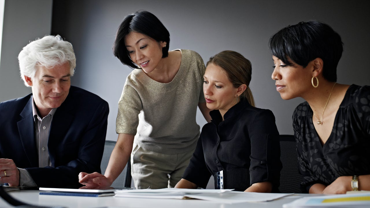 A businesswoman leans over a table, looking over papers with her diverse staff