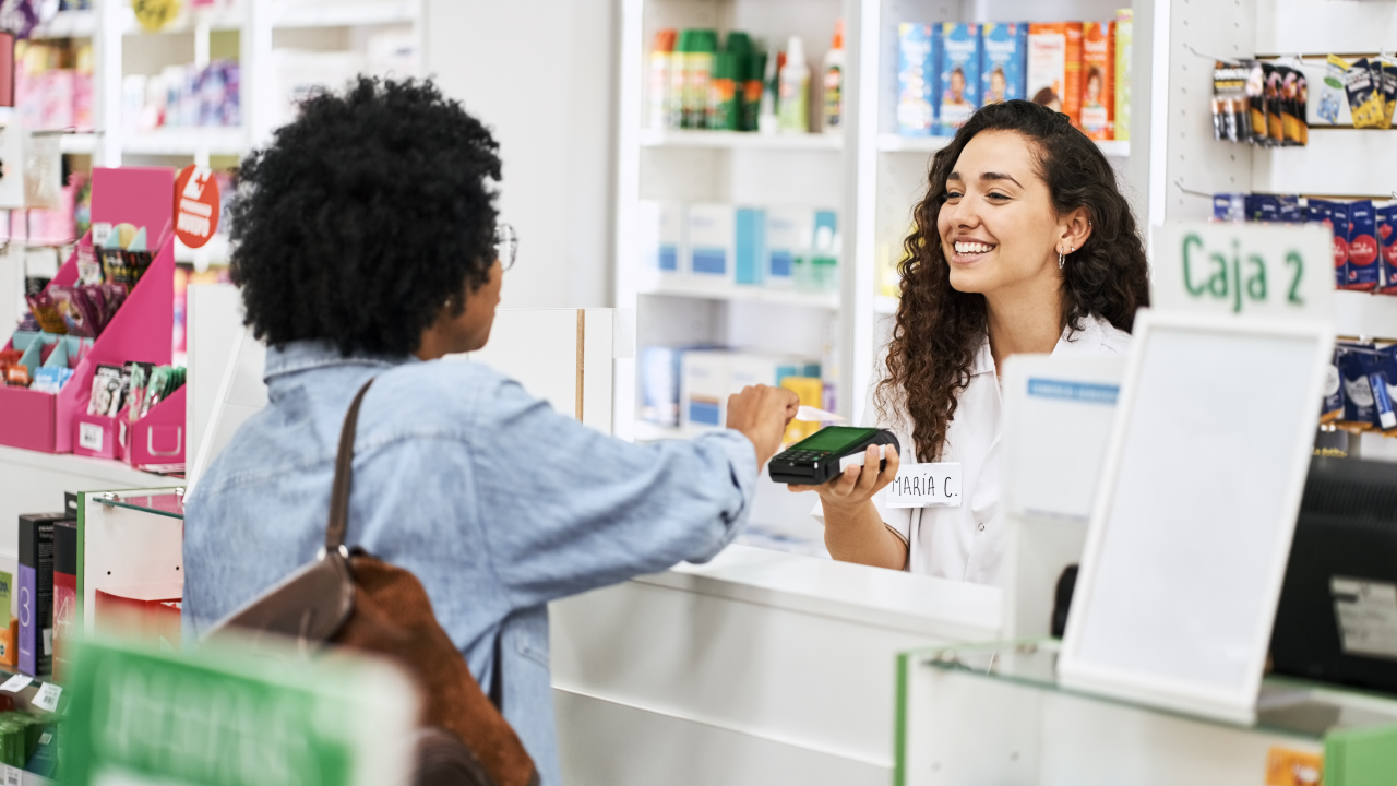 Female customer doing nfc payment with her credit card at pharmacy. Smiling cashier holding credit card reader at drug store checkout counter.