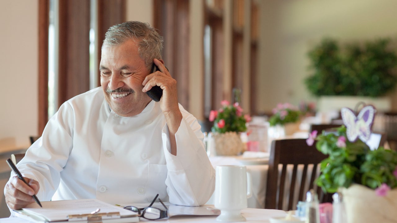 A chef sits at one of his tables in his restaurant, talking on his phone and looking over papers.