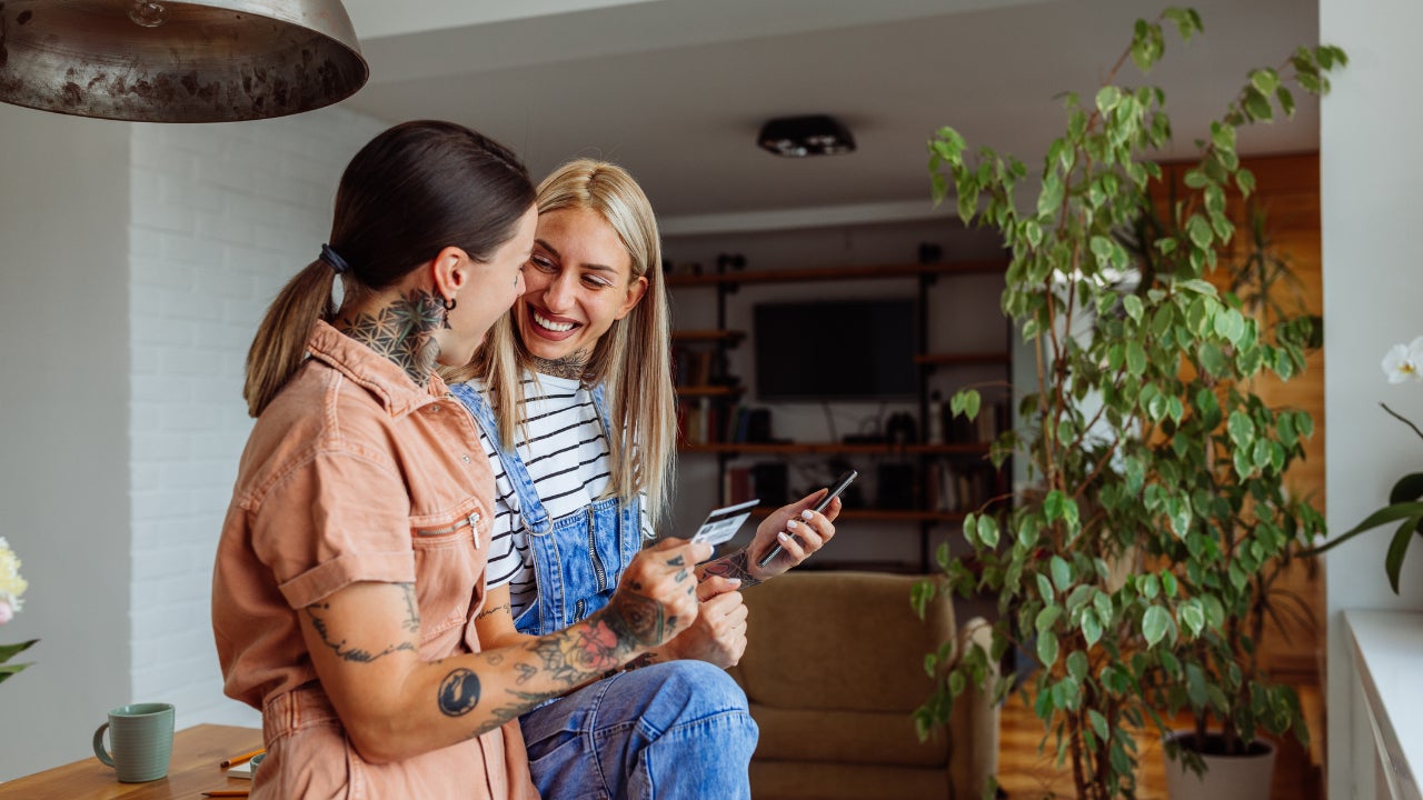 Two women smile at one another. One sits on a table holding a phone, while the other stands holding a credit card.