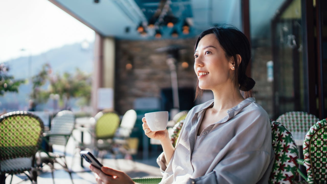 Cheerful young Asian woman using smartphone while sitting at a sidewalk cafe.