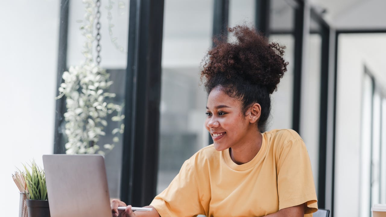 An attractive and happy millennial businesswoman is sitting on a sofa in her living room, working with a laptop computer at home.