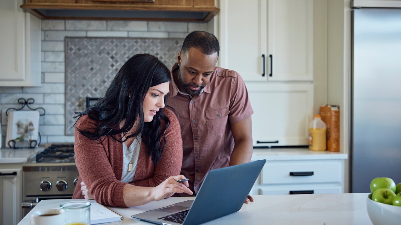 Woman and man sit at kitchen counter with breakfast working with pen, paper and laptop