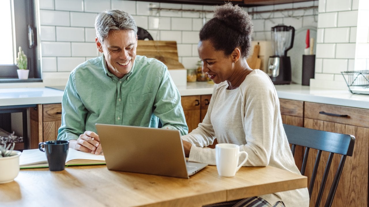 A white man and Black woman sit together at a kitchen table while working on a laptop.