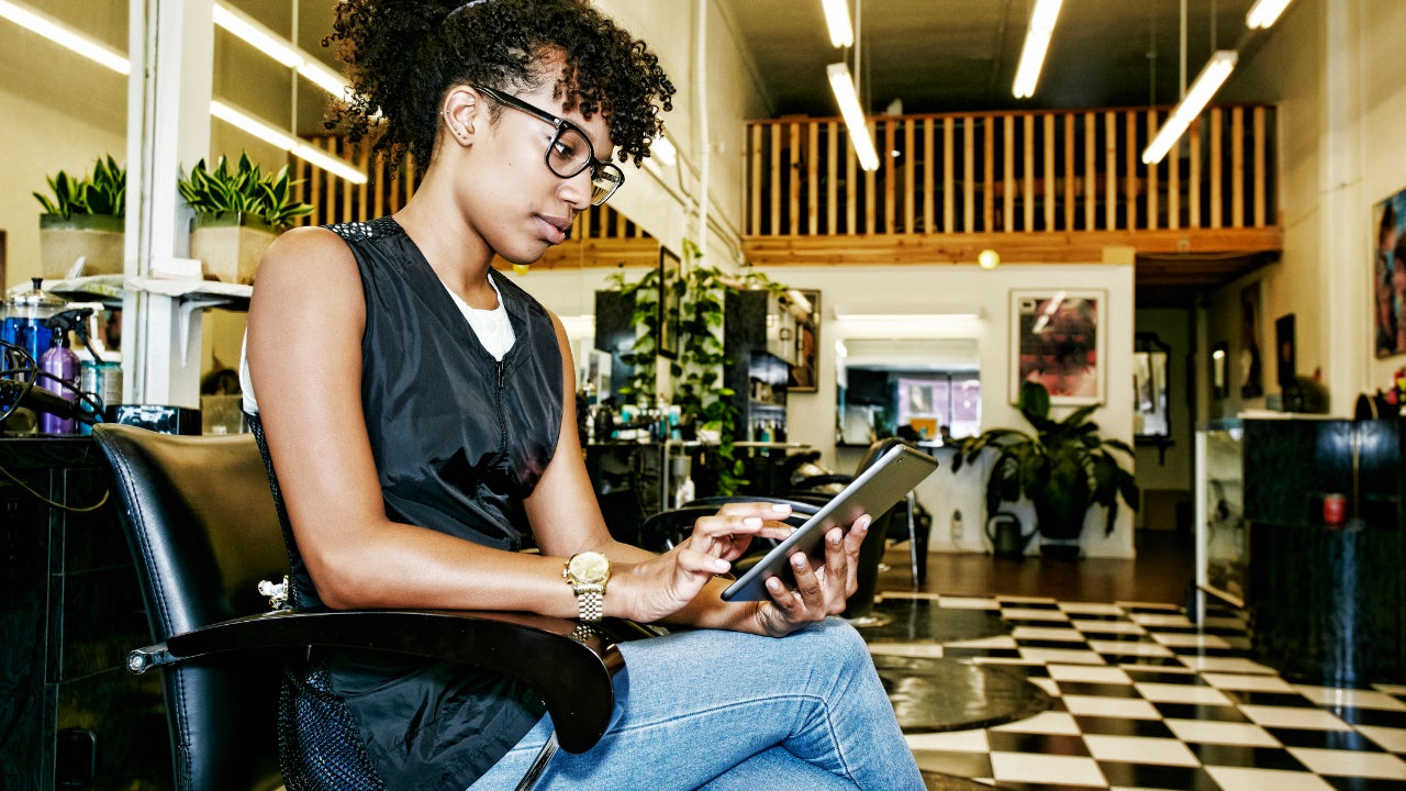 A young woman sits in the chair at her empty salon, looking at her tablet.