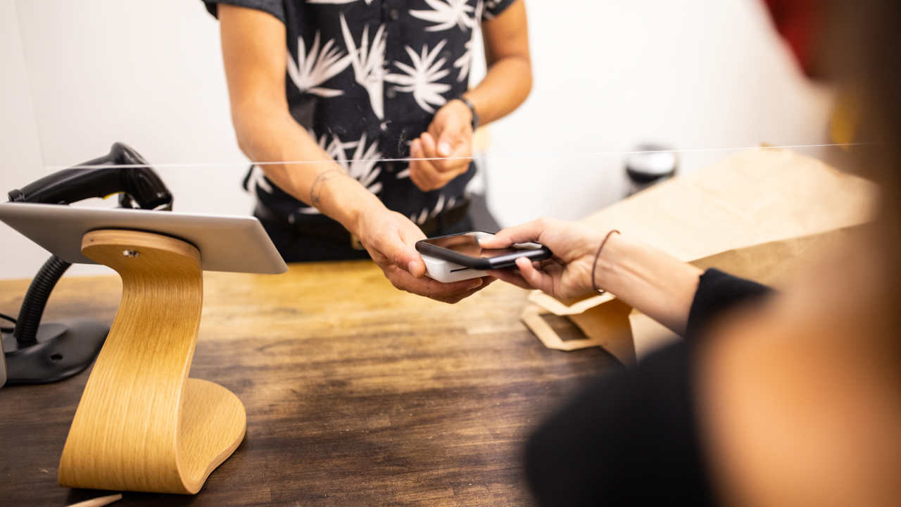 Close-up of a female customer in boutique making contactless mobile payment for her purchase. Customer holding cell phone over credit card reader at checkout counter with a sneeze guard in boutique.