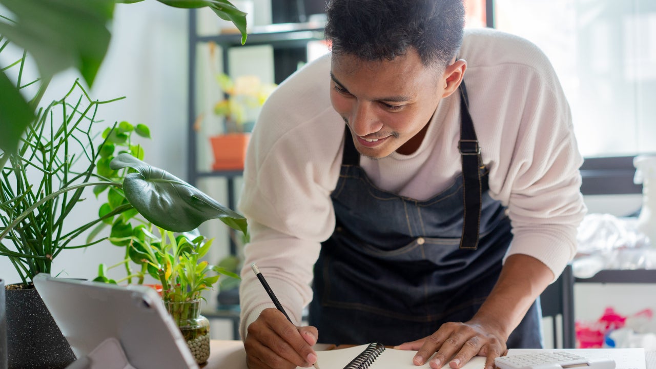 A smiling young business owner leans over his desk, taking notes while looking at a tablet.