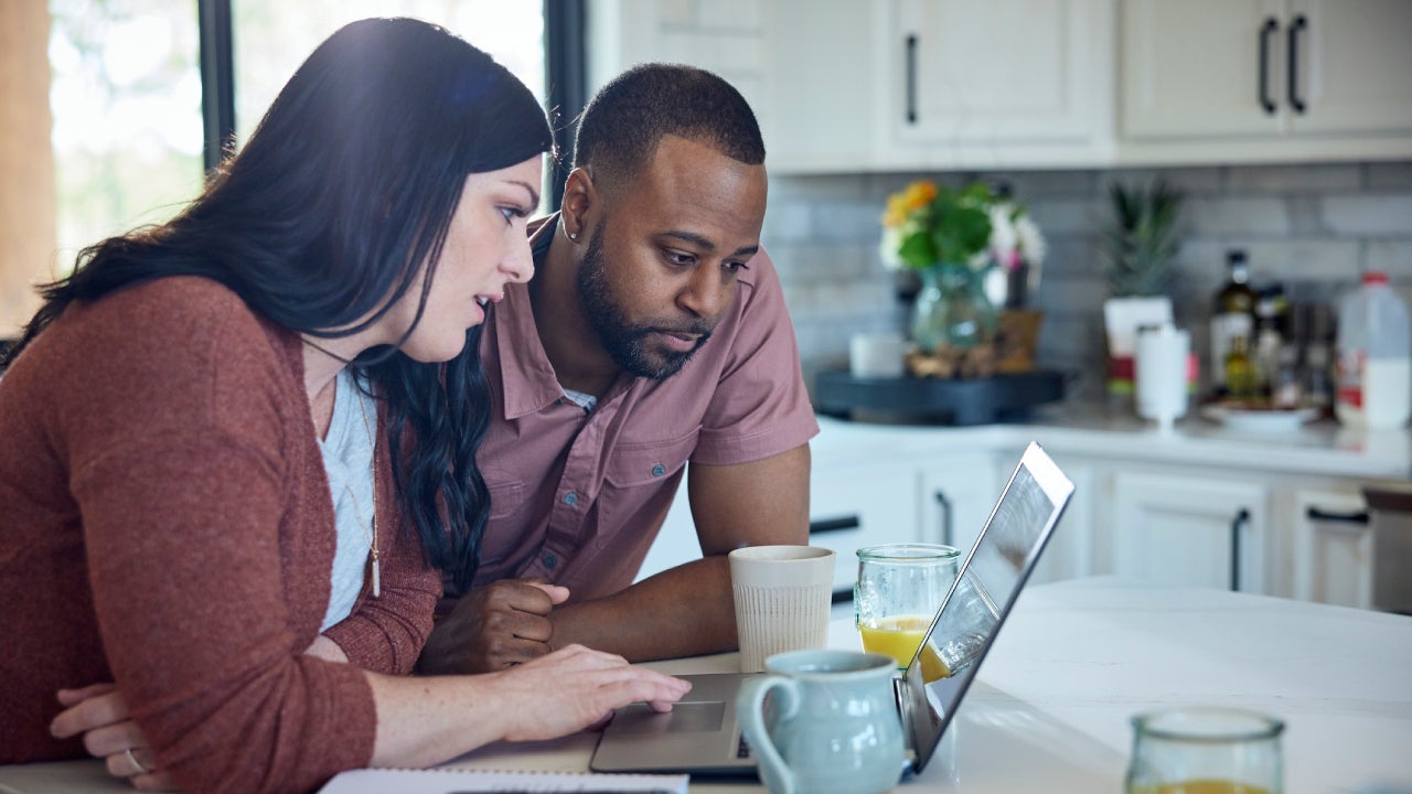 Married couple sit at kitchen counter with breakfast working with pen, paper and laptop