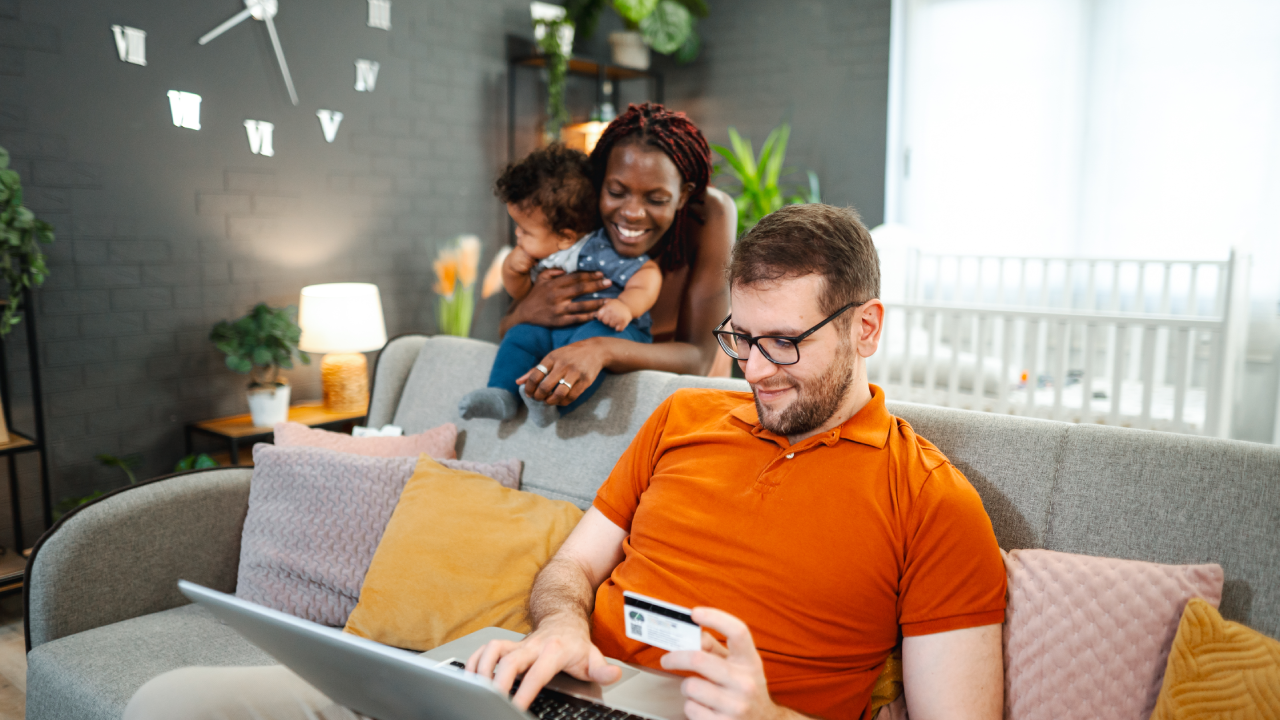 Husband sitting on the couch online shopping while his wife is holding baby son behind