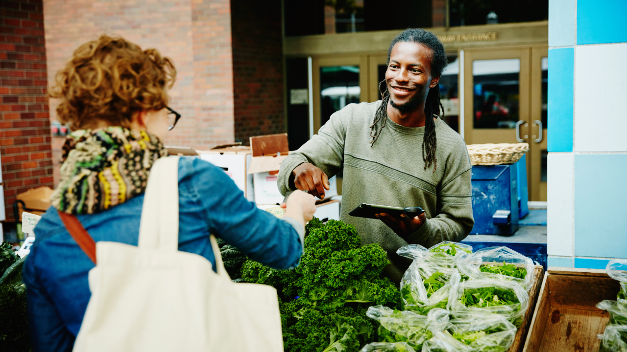 Smiling organic farmer taking credit card from customer to run transaction on digital tablet at farmers market