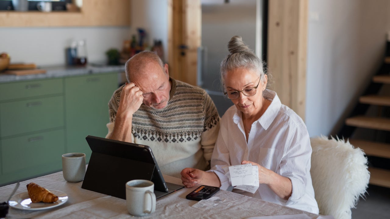 Senior couple sitting at the kitchen table with croissant and cups of tea, looking at digital tablet and recalculating their expenses.