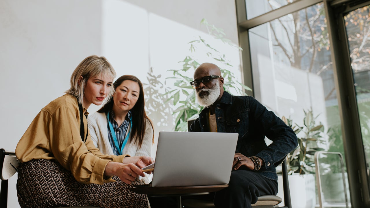 Three people sit around a laptop. One woman is presenting to the group.